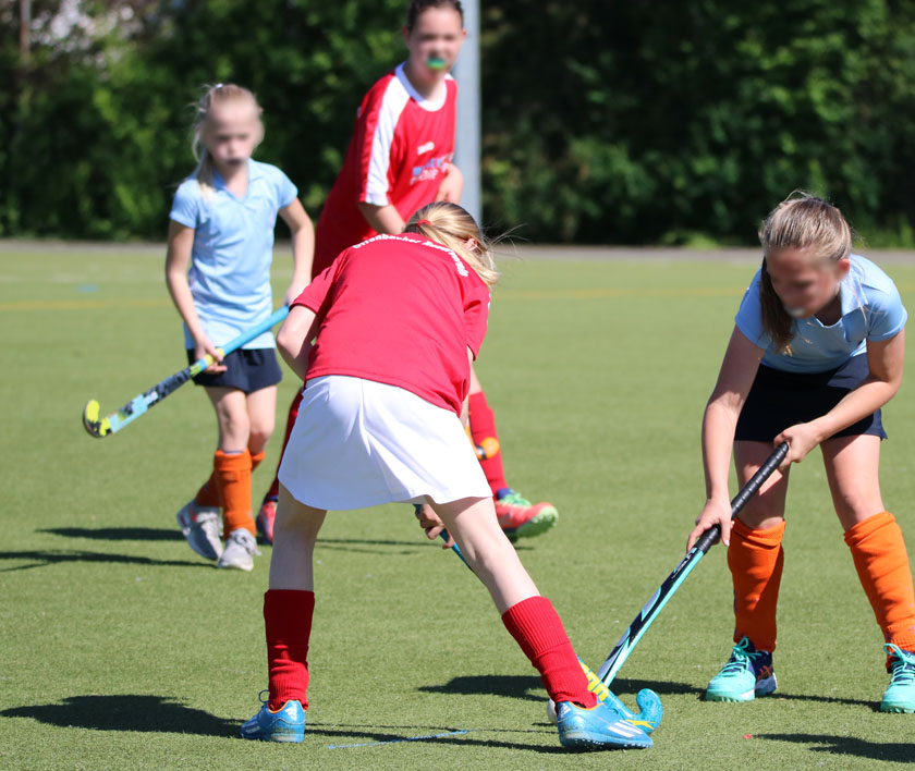 Vier Mädchen spielen Hockey auf einem grünen Kunstrasenfeld. Sonniger Tag, teils bewaldete Umgebung.