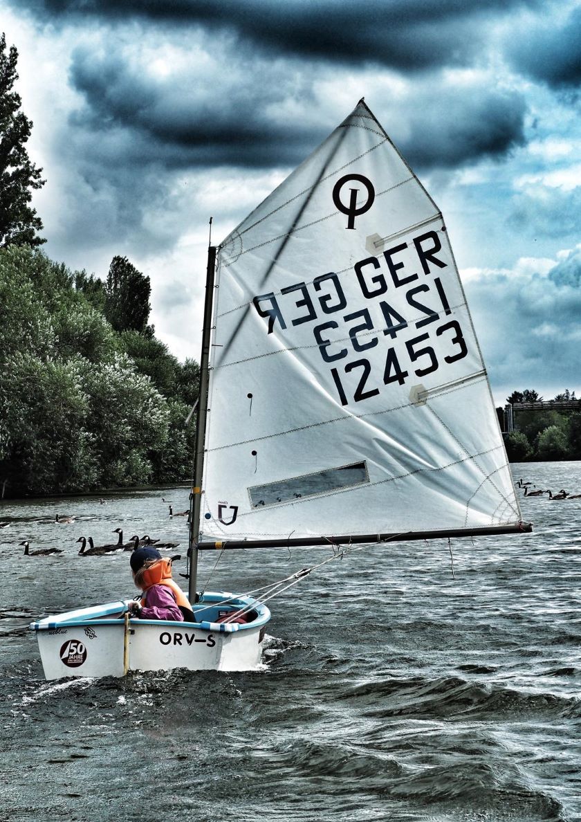 Ein Kind segelt in einem kleinen Boot auf einem Gewässer, umgeben von Enten und bewölktem Himmel.