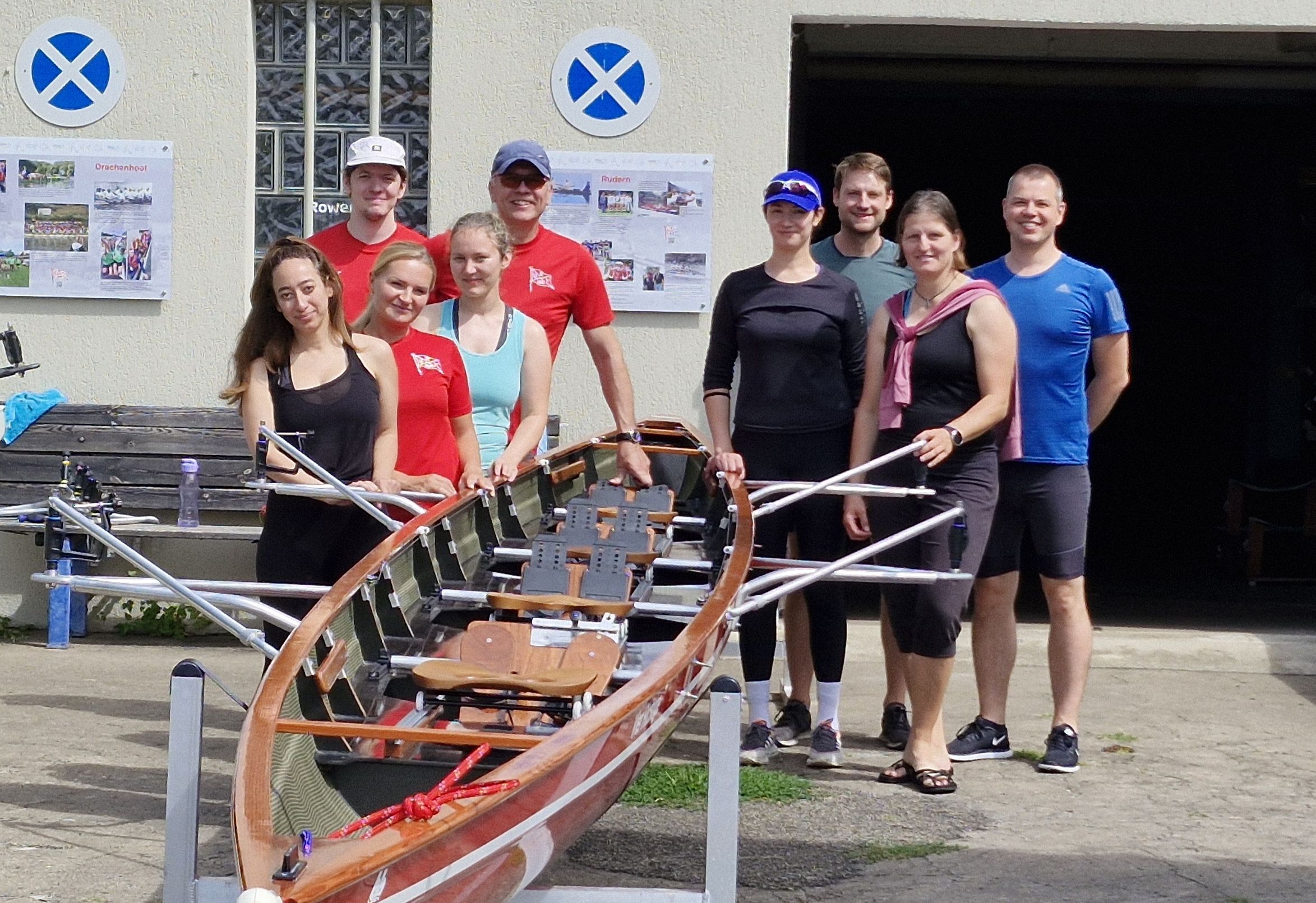 Neun Personen stehen um ein Ruderboot, im Hintergrund ein Vereinsheim. Alle sind in Sportswear.