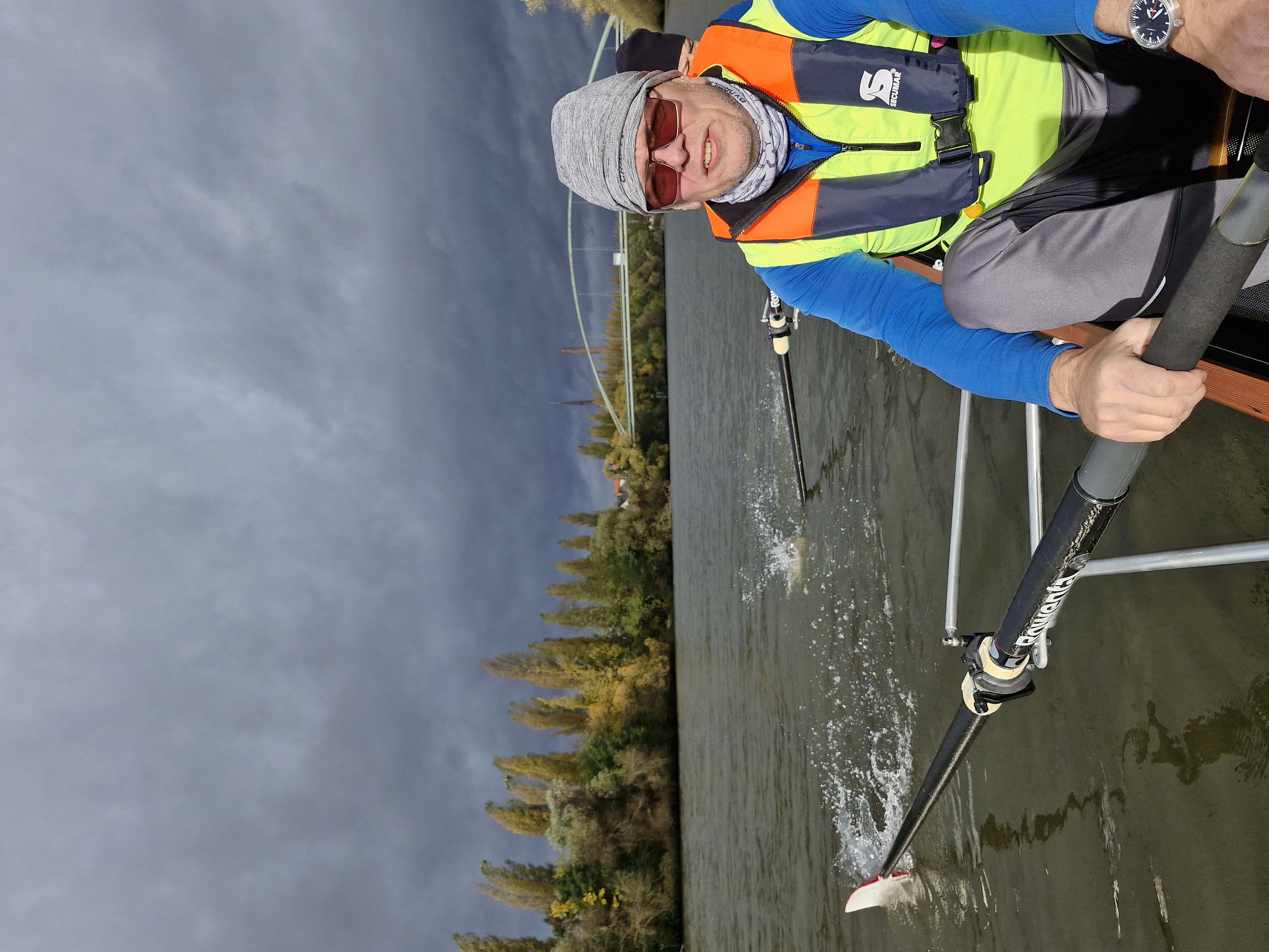 Ein Mann rudert auf einem Gewässer, umgeben von Bäumen und bewölktem Himmel.