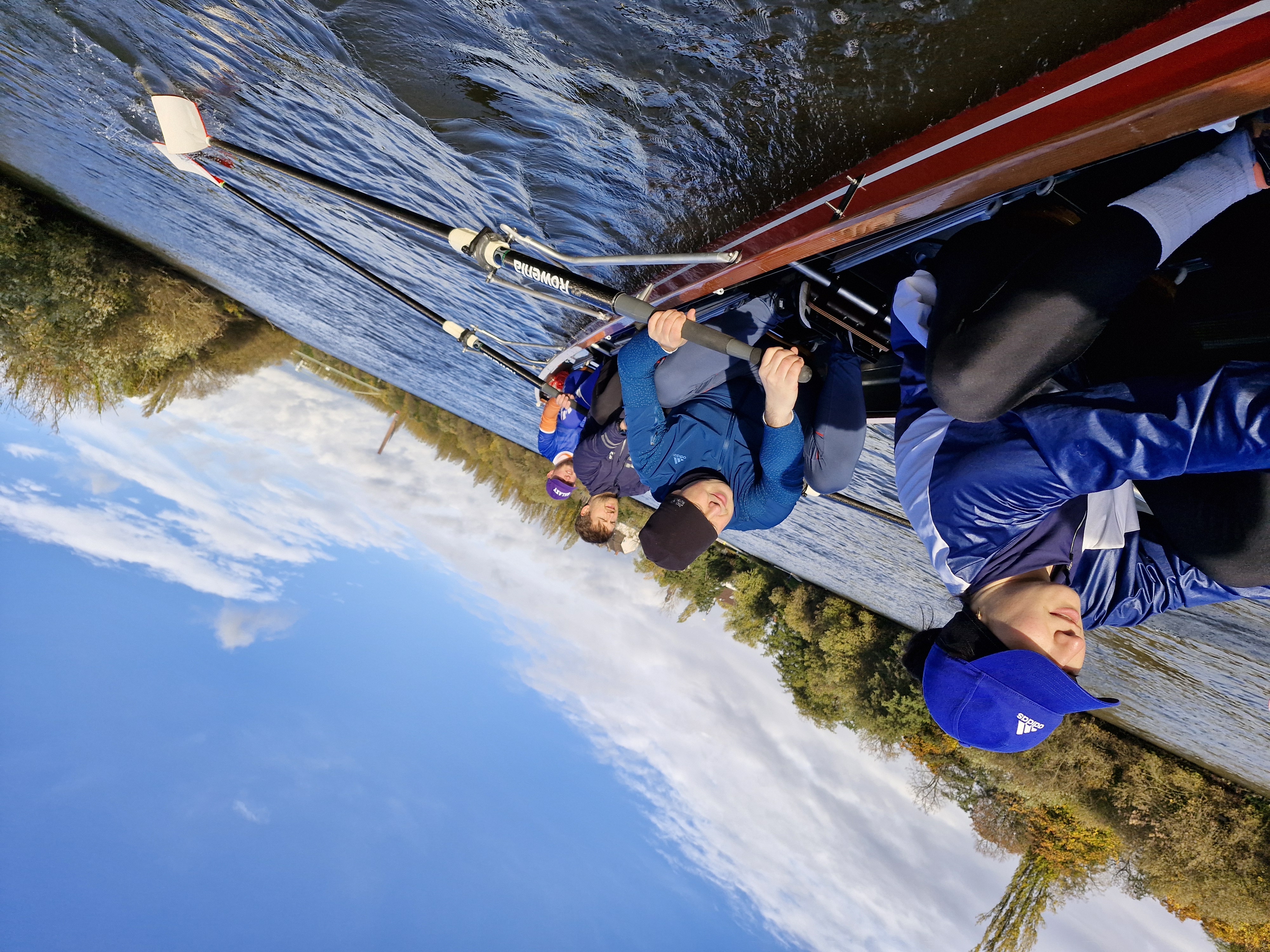 Fünf Personen rudern gemeinsam in einem Boot auf einem Gewässer, umgeben von Bäumen und Himmel.