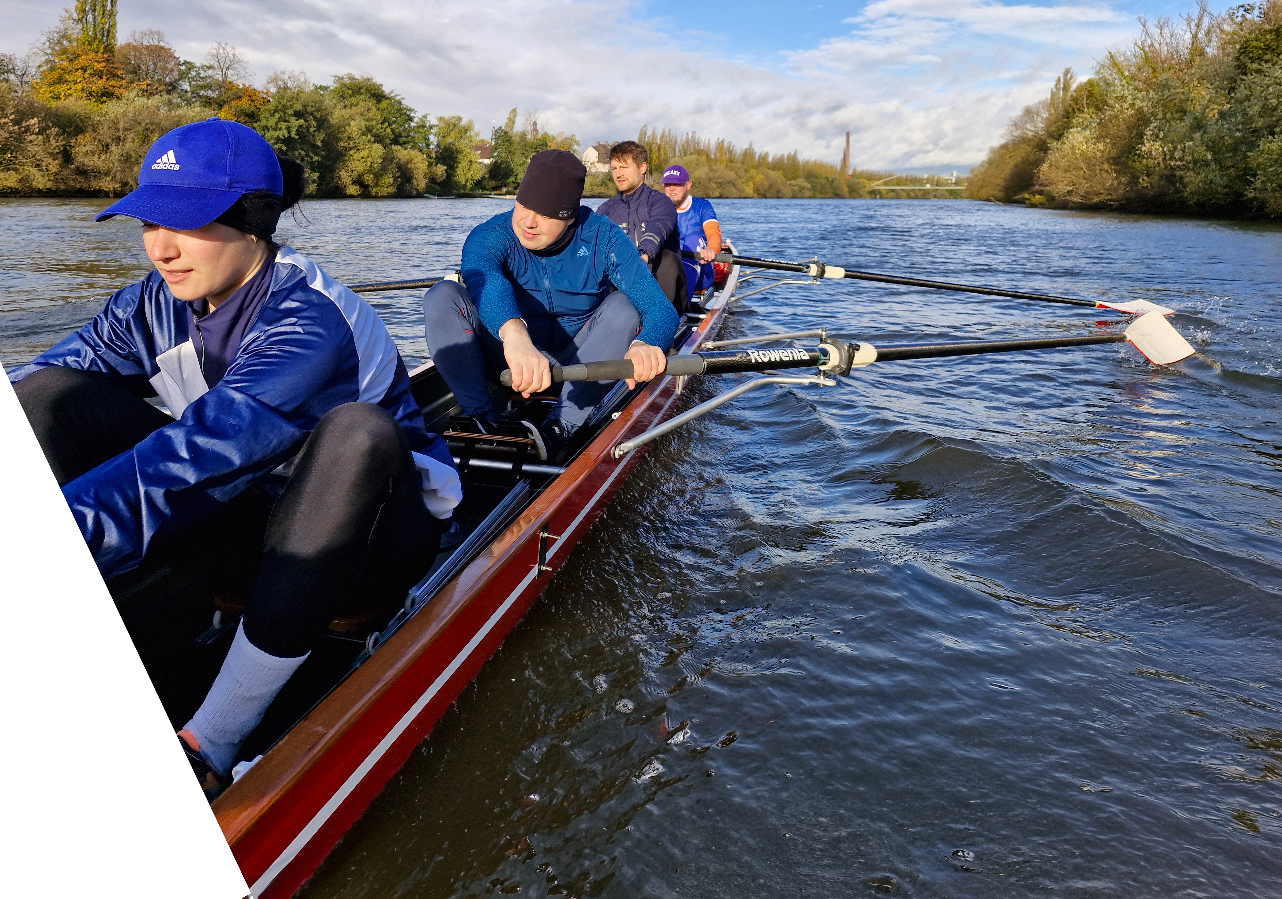 Fünf Ruderer in einem Boot auf einem Fluss, umgeben von Bäumen und blauem Himmel.