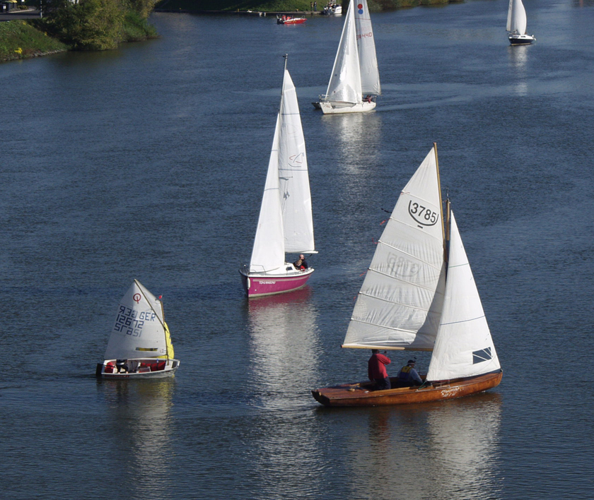 Drei Segelboote mit insgesamt vier Personen auf einem ruhigen Gewässer, umgeben von Bäumen.