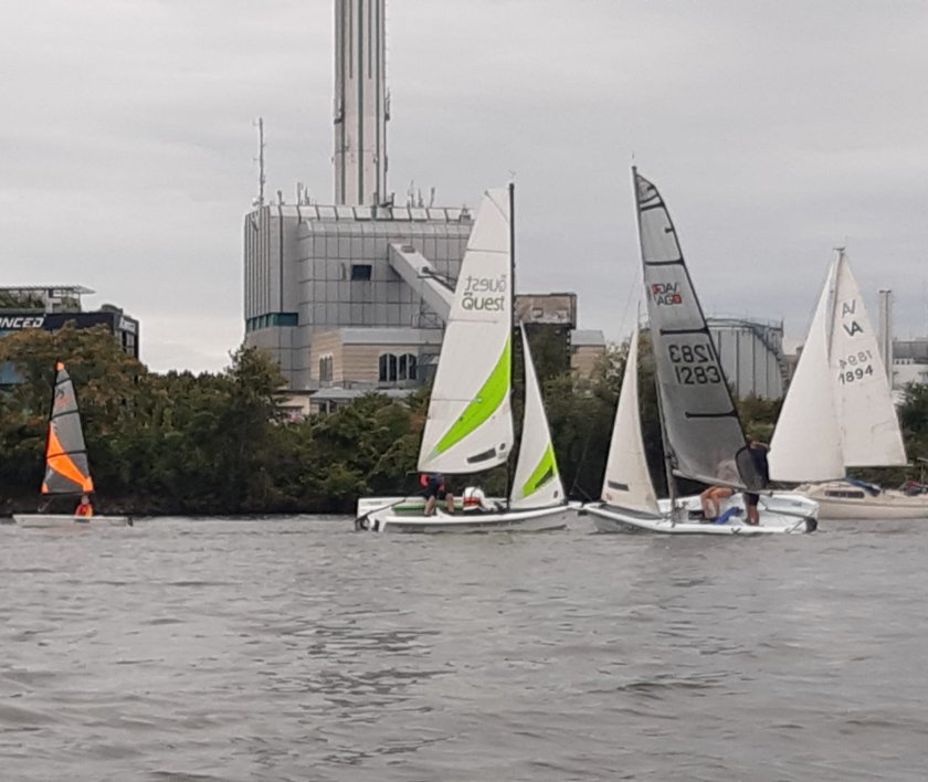 Segelboote auf einem Gewässer mit modernen Gebäuden im Hintergrund, sanfte Wellen, bewölkter Himmel.