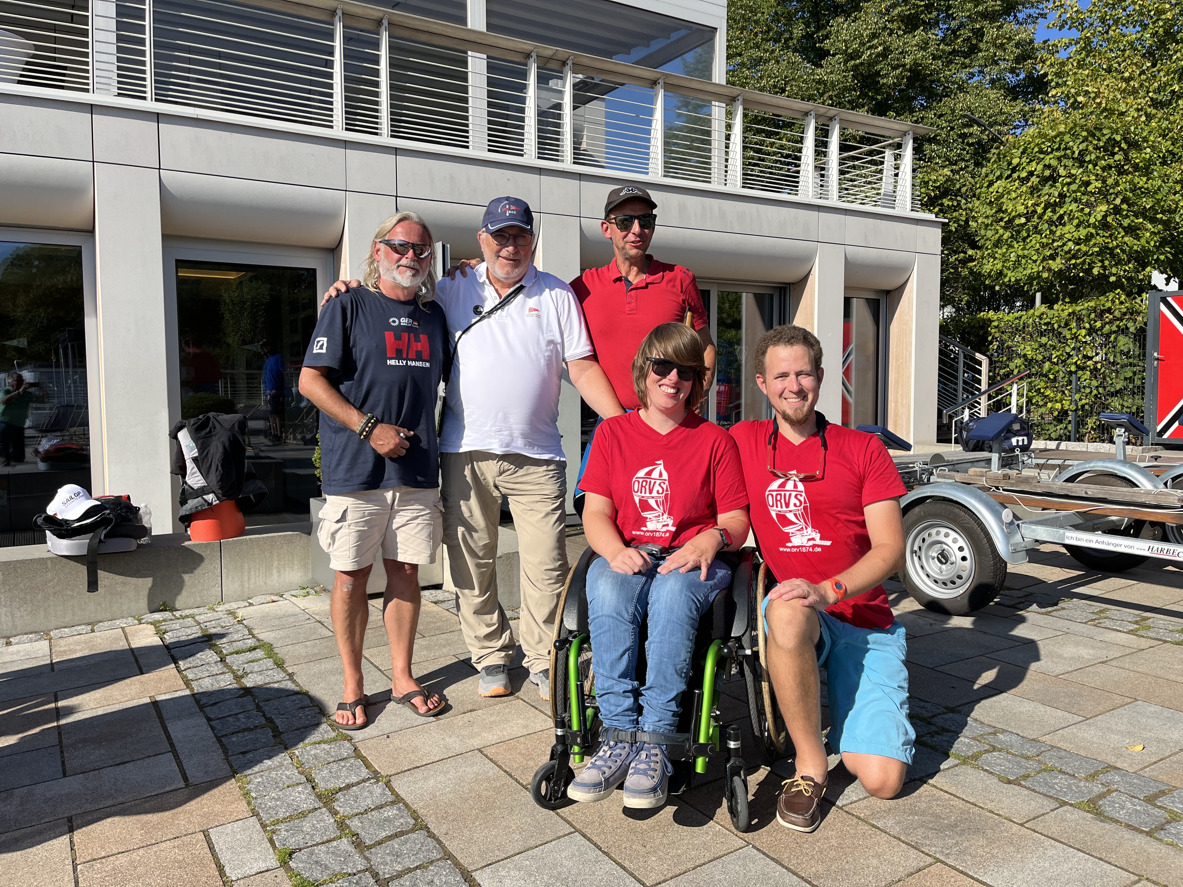 Fünf Personen in Vereinskleidung stehen und sitzen vor einem Vereinsgebäude am Wasser, lächeln freundlich.