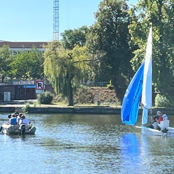 Zwei Boote auf einem Gewässer, ein Segelboot mit zwei Personen und ein Ruderboot mit mehreren Passagieren.