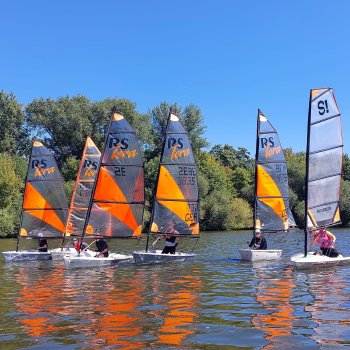 Sechs Segelboote mit orange-schwarz-gestreiften Segeln auf einem Gewässer bei sonnigem Wetter.