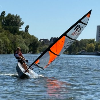 Zwei Personen segeln auf einem Sportboot auf einem Gewässer unter blauem Himmel.