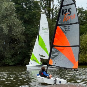 Kind mit orangefarbenem Segelboot segelt auf einem Gewässer, im Hintergrund ein weiteres Boot mit grünem Segel…