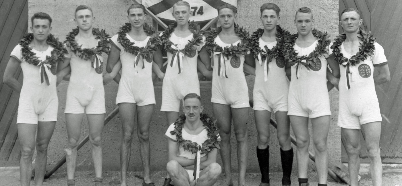 Acht junge Männer in Sportkleidung mit Lorbeerkränzen, stehen vor einer Wand mit Flagge.