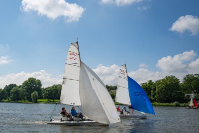 Zwei Segelboote mit je mehreren Personen segeln auf einem Gewässer unter blauem Himmel.