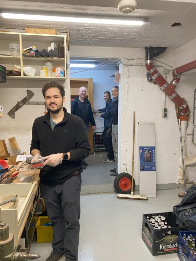 Vier Männer in einem Werkstattbereich, einer hält ein Werkzeug und blickt zur Kamera.