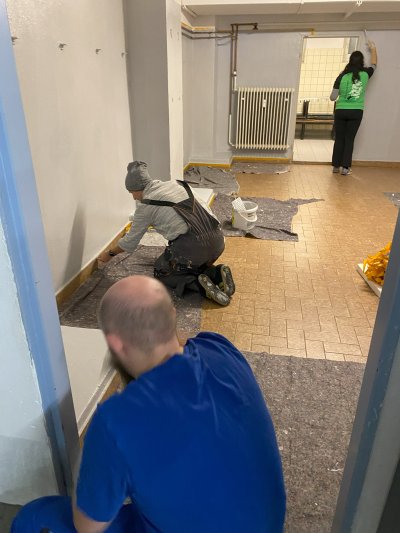 Drei Personen streichen Wände in einem Raum, eine sitzt am Boden, zwei arbeiten an den Wänden. Drei Personen streichen Wände in einem Raum, eine sitzt am Boden, zwei arbeiten an den Wänden.