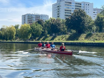 Fünf Personen rudern in einem Boot auf einem ruhigen Gewässer, umgeben von Wohngebäuden und Bäumen.