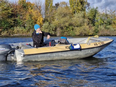 Ein Mann in blauer Mütze sitzt in einem kleinen Boot auf einem Gewässer, umgeben von Bäumen.