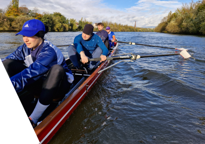 Fünf Ruderer in einem Boot auf einem Fluss, umgeben von Bäumen und blauem Himmel.