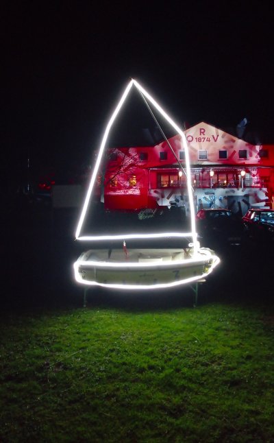 Beleuchtetes Segelboot im Vordergrund, im Hintergrund ein Gebäude mit roter Fassade und Fenstern. Beleuchtetes Segelboot im Vordergrund, im Hintergrund ein Gebäude mit roter Fassade und Fenstern.