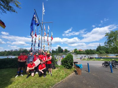 Gruppe von etwa 15 Personen in roten T-Shirts posiert am Wasser, im Hintergrund Boote und Flaggen.